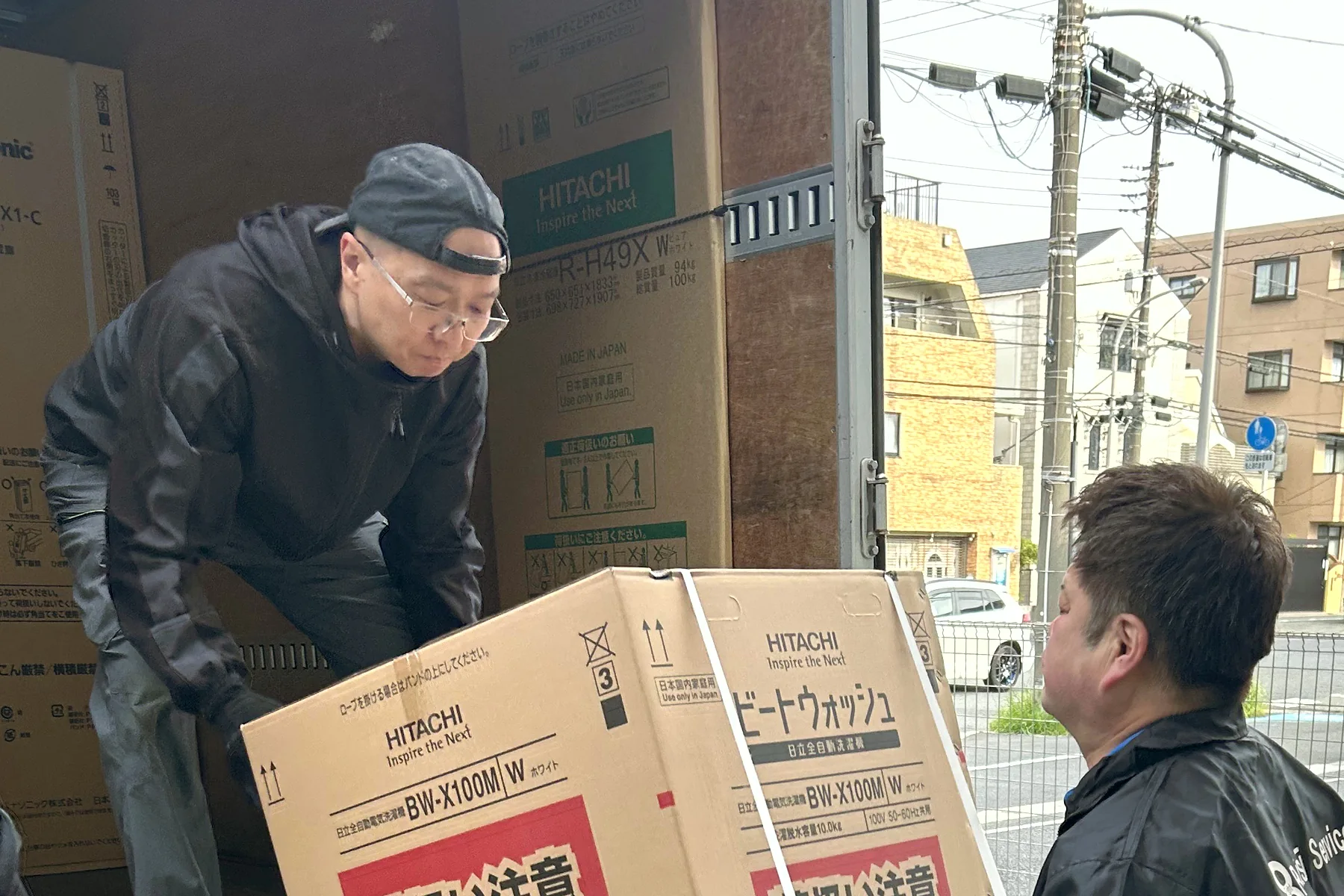 住宅街で、洗濯機の箱を2人で受け渡すところ